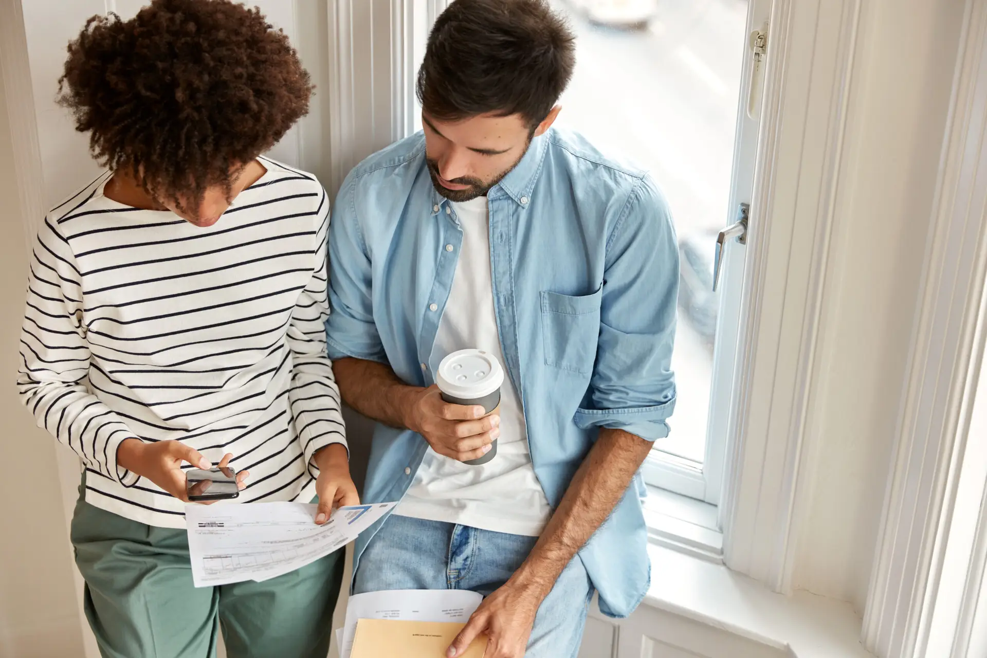 Two people sit by a bright window; one holds documents and a smartphone, while the other has a coffee cup and folder. They review papers together, possibly discussing home inspection price in Texas or the Super Inspector FlexFund payment options.