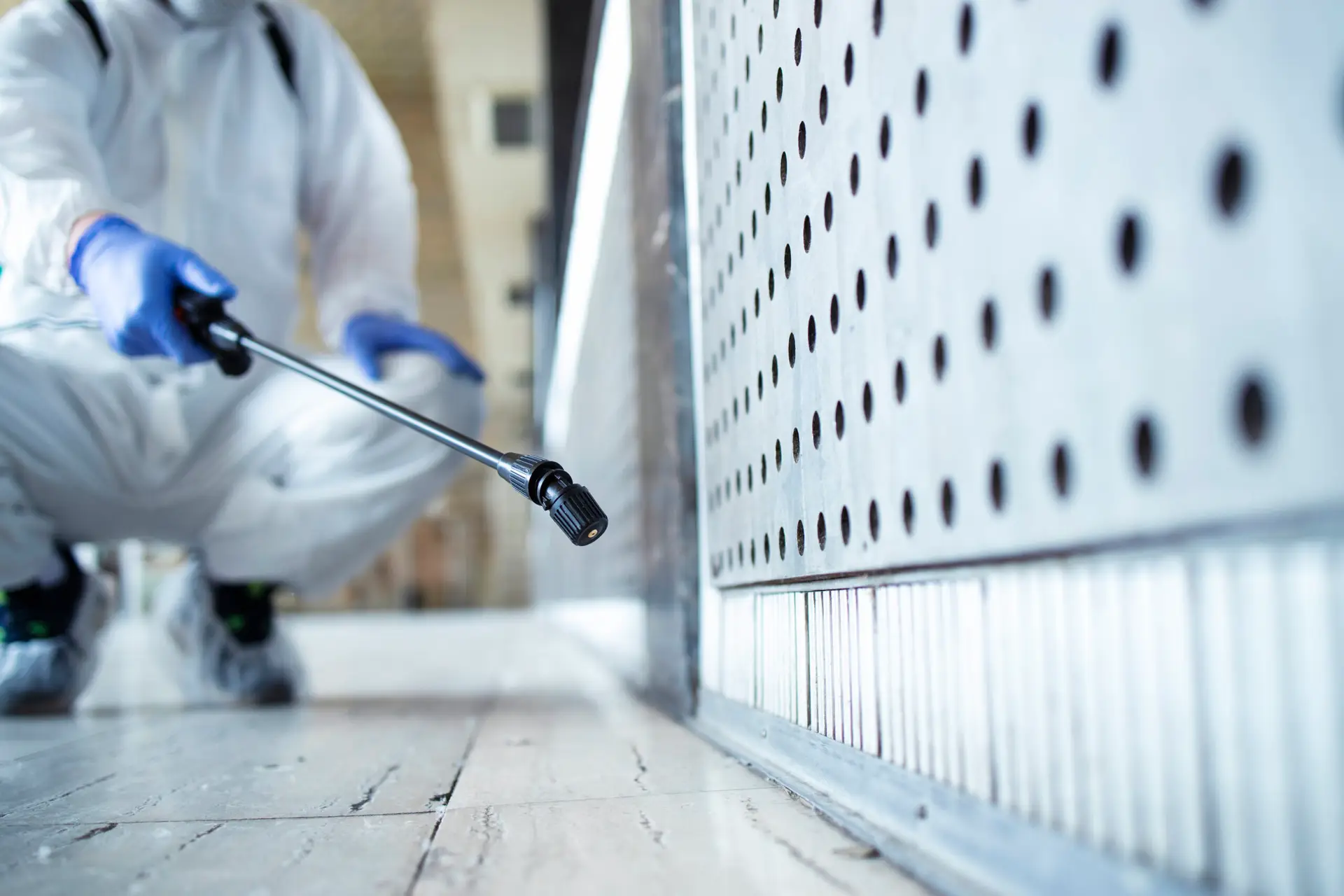 A person in protective gear and blue gloves crouches while using a spray wand to apply chemicals along a wall base, likely performing pest management or disinfection in an indoor space.