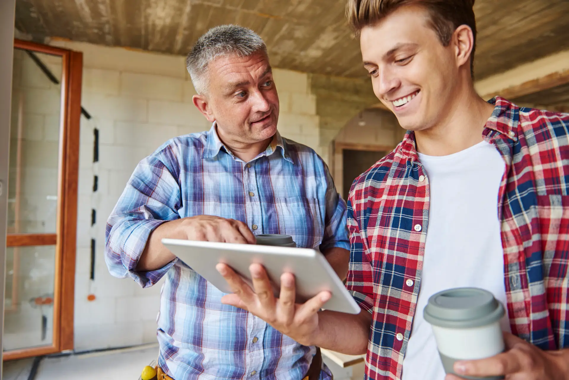 Two men in casual plaid shirts stand in a partially constructed room, smiling and looking at a tablet. Representing a home inspection company, one holds a reusable coffee cup while the other points at the screen, suggesting collaboration on Home Inspections in Frisco.